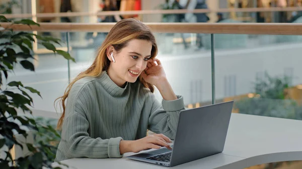 Side view young beautiful female freelancer girl business woman in wireless headphones with laptop in public shopping center company office looking at laptop smiling happy video call reads good news — стоковое фото