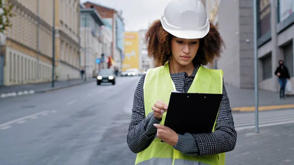 Retrato joven afroamericano hermosa mujer ingeniera con chaleco de seguridad y sombrero de pie en la ciudad cerca de datos de escritura de tráfico por carretera. Mujer profesional que trabaja en la fabricación logística al aire libre —  Fotos de Stock