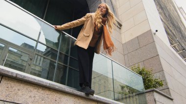Alt manzara, şık güzel kız ofis binasının yanındaki terasta duruyor. Dalgın kadın öne eğildi ve kafasını kaldırdı. Kafkasyalı iş kadını elinde balkonla dışarıda dinleniyor.