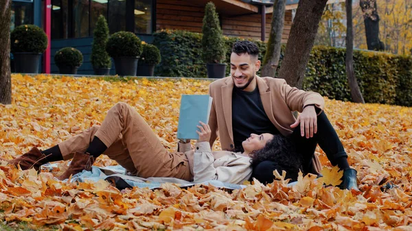 Fecha al aire libre joven barbudo árabe chico sinceramente hablando con la mujer amada hombre asiente aprobando hermosa chica acostada en cuadros en hojas amarillas libro de lectura feliz pareja sonriendo juntos en el parque de otoño — Foto de Stock