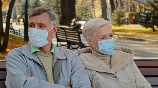 Elderly family couple in medical masks sit in autumn park mature man turns away resentfully crossing arms unpleasant conversation outdoors misunderstanding spouses angry and frustrated after quarrel — Foto Stock
