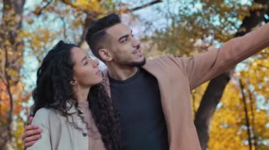 Happy hispanic young couple hugs in autumn park joyfully looking into distance enamored guy and girl cute communicate woman looks upward showing on sky outdoors man point finger side thinking dreamily