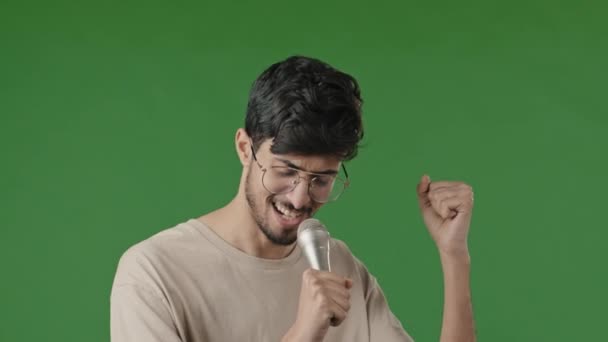 Árabe macho en verde fondo en estudio cantando karaoke uso micrófono primer plano alegre joven con gafas activamente bailando feliz sonriente juguetón chico divertirse mirando cámara disfrutar ritmo de canción — Vídeos de Stock