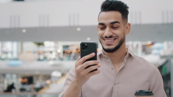 Close-up businessman walking in mall holding mobile phone hispanic guy smiling drinking coffee reading good news using device young man chatting in social media enjoying weekend male make online order — Stockvideo