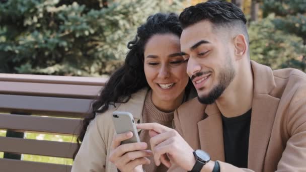 Young hispanic married couple discussing online order happy man woman smiling browsing websites using telephone sit on bench in park family talking swipe virtual electronic catalogue pages on mobile — Vídeo de Stock