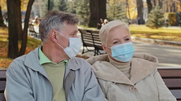 Elderly family couple in medical masks sit in autumn park mature man turns away resentfully crossing arms unpleasant conversation outdoors misunderstanding spouses angry and frustrated after quarrel — Wideo stockowe