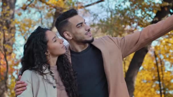 Happy hispanic young couple hugs in autumn park joyfully looking into distance enamored guy and girl cute communicate woman looks upward showing on sky outdoors man point finger side thinking dreamily — Video