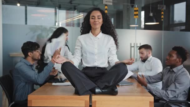 Young hispanic woman sitting on table in lotus position female leader meditating in office businesswoman feels calmness and serenity annoyed angry colleagues throw documents patience workplace concept — Video Stock