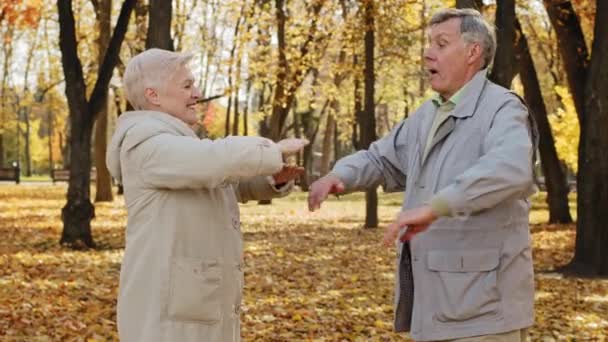 Joyful elderly married couple doing exercises outdoors positive old grandpa and grandma smiling happily caucasian active spouses aged in autumn park together training muscles healthy lifestyle concept — Wideo stockowe