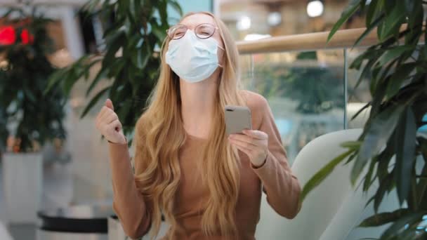 Happy young girl in medical mask sit in mall got sms with good news on smartphone joyfully dancing celebrating win online lottery caucasian woman holding telephone rejoice success make victory gesture — Vídeo de Stock