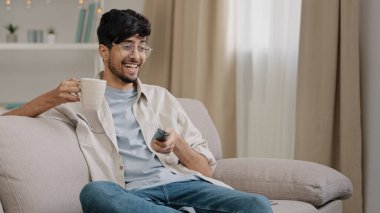 Arab man bearded guy with glasses resting at home sitting on couch drinking tea coffee watching funny movie championship sports game TV rejoices at victory of favorite team laughing fun sincerely