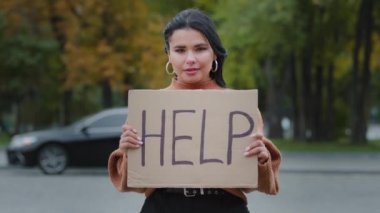 Close-up sad young woman stands outdoors near roadway holding cardboard sign with words HELP upset hispanic girl searching for job experiencing financial crisis problems of dismissal unemployment