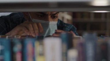 Close-up hispanic guy student in protective medical mask looking for right book on bookshelf in university library or bookstore serious young man search textbook for homework essay college education