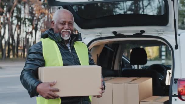 Starší africký americký usměvavý muž drží lepenkové krabice stojí v blízkosti otevřeného kufru auta šťastný starší muž kurýr venku v pracovním oblečení přijímá zboží kutil nakladač zatížení krabice dodávky — Stock video