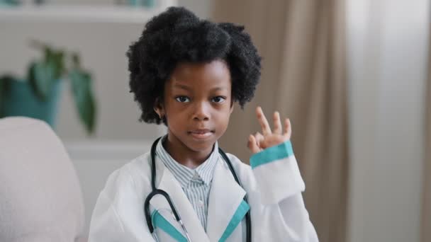 Крупним планом афро-американська дівчинка, що стоїть в медичному одязі, грає прикидаючись лікарем чарівною дитиною, дивлячись на камеру посміхається показує добре жест ознаки без проблем концепція майбутніх професій — стокове відео