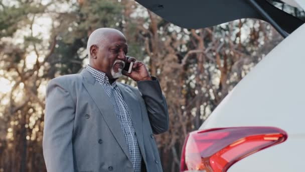 Close-up african american businessman stands outdoors near open trunk of car elderly man entrepreneur talking on mobile telephone business conversation using smartphone mature male answers phone call — Vídeo de Stock