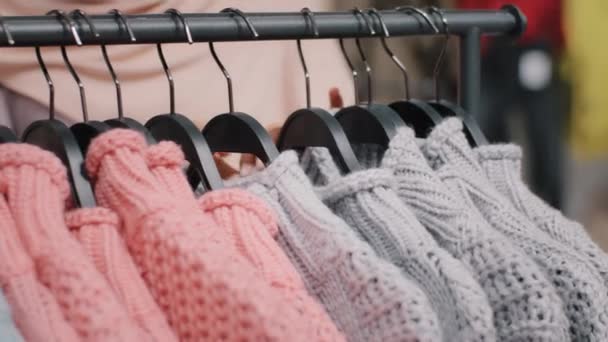 Магазин одягу крупним планом вішалка для одягу з яскравими кольоровими светрами жіночі руки невпізнаваної дівчини вибирає одяг в торговому центрі модний дизайн якісний матеріал дисконт продаж — стокове відео