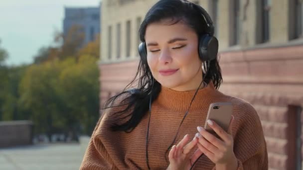 Mujer joven despreocupada al aire libre en la ciudad escuchando música en los auriculares desde el teléfono inteligente linda chica hispana feliz bailando a la canción favorita disfrutar del sonido de audio en línea utilizando la aplicación en el teléfono móvil canta sonrisas — Vídeo de stock