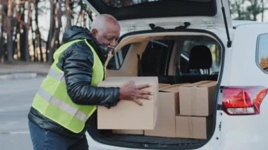 Close-up mature delivery service worker unloading cardboard boxes from trunk of car elderly african american courier loader delivering goods handyman smiling in work clothes accepts shipping outdoors