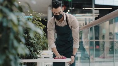 Young guy waiter wipes table in cafe food service worker in medical mask does cleaning before opening of restaurant student works part-time hispanic man finishes shift covid 19 coronavirus epidemic
