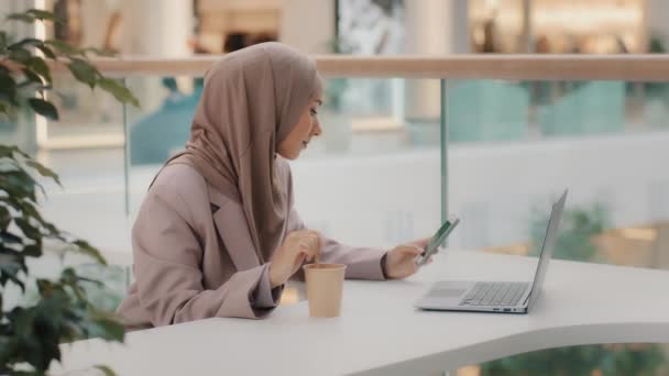 オフィスに座っているヒジャーブの若いイスラム教徒のビジネスマンの女性は、紙コップレジャーアラブの女の子で電話攪拌コーヒーを使用していますスマートフォンで楽しい電子メールを読んで思慮深く恥ずかしそうに笑顔受信した日付招待状 — ストック動画