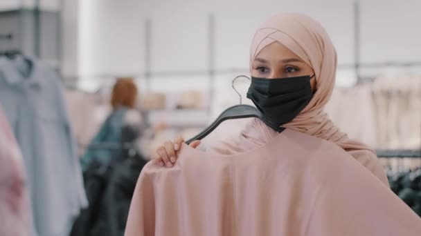 Cabeza joven musulmana mujer en máscara de protección médica durante la cuarentena chica cliente en hijab en la tienda de ropa tratando de vestirse mirando en espejo cliente femenino selecciona productos en la venta de ropa de descuento — Vídeo de stock