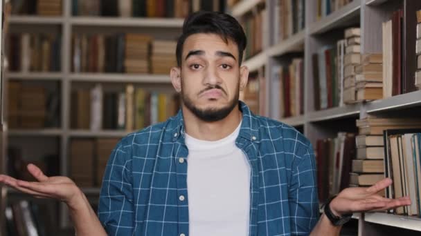 Close-up young upset man standing indoors in university library hispanic student misunderstood puzzled shrugs shoulders doesnt know what to answer doubt insecure guy looking at camera with sadness — Stok video