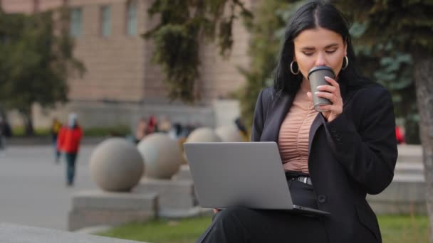 Close-up young businesswoman sitting in city outdoors girl student freelancer working studying on laptop drinks coffee tea from disposable cup hispanic cute woman browses online shopping on internet — Stockvideo