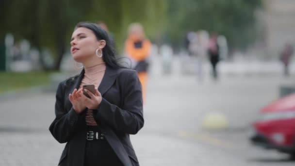 Close-up businesswoman stands outdoors in city near roadway holding mobile phone looking around focused hispanic girl trying find building using electronic map geolocation application in smartphone — ストック動画