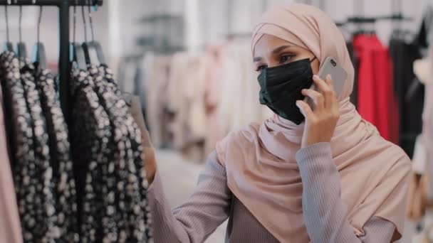 Close-up young muslim woman in medical protective mask talking on cell phone with friends uses smartphone arab girl making purchases chooses outfit tries on blouse buying clothes in clothing store — стоковое видео