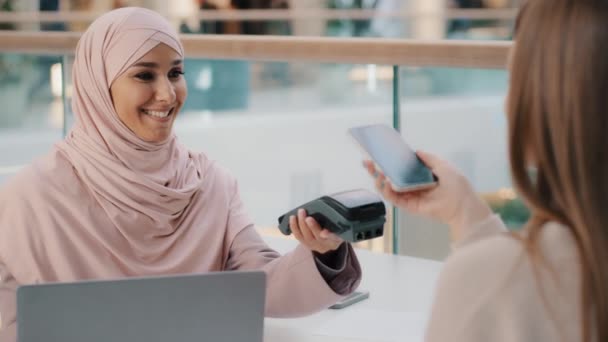 Friendly muslim woman in hijab seller agent offers pay for service through bank terminal girl shopper consumer pays for purchase using contactless payment technology on smartphone client uses phone — ストック動画