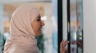 Young happy arab woman in hijab stands near coffee machine orders drink smiling muslim girl making order on self-service automaton pays for service using contactless payment technology on smartphone