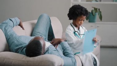 Ciddi çocuk, doktor gibi davranır. Tablete semptomlar yazar. Babası kanepeye uzanır.