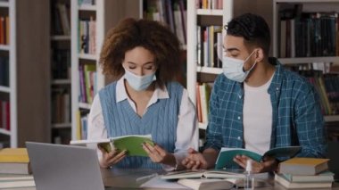 Students in medical masks sitting in university library communicate with teacher on webcam via video call using laptop doing homework hispanic guy help african american girl understand information