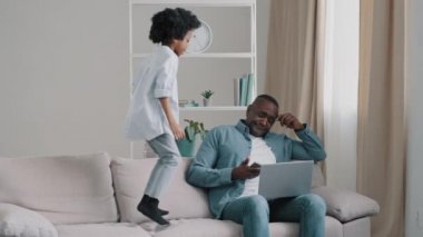 Küçük kız kanepede hoplayıp zıplıyor, çocuk yüzünden kızgın, kötü davranışlar gösteriyor kız babasına karışıyor erkek akıllı telefondan saate bakıyor gürültücü çocuk yüzünden işi bitirecek zamanı olmuyor.