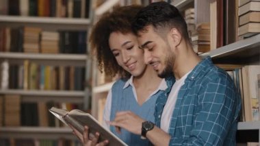 Young couple students standing in university library laughing reading funny story hispanic guy with african american girl holding book in hands studying looking at textbook school college education