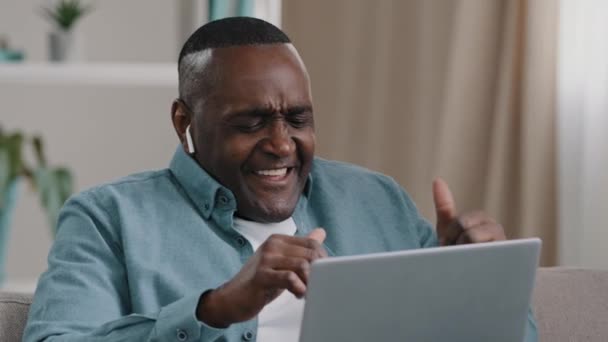 African american businessman freelancer distracted from work by laptop happy man sitting home on sofa listening to music in wireless headphonesenjoys favorite song dancing to energetic rhythm of audio — Video