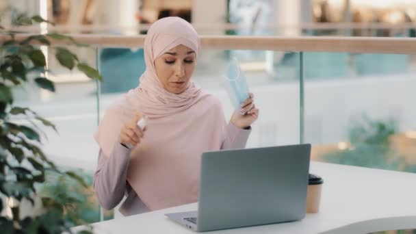 Забавна арабська жінка, сидячи в громадському місці, використовує антисептичні зморшки навколо в повітрі на обкладинках ноутбука обличчя з медичною маскою мусульманська дівчина боїться мікробів, використовуючи дезінфікуючий спиртний спрей запобігає зараженню — стокове відео