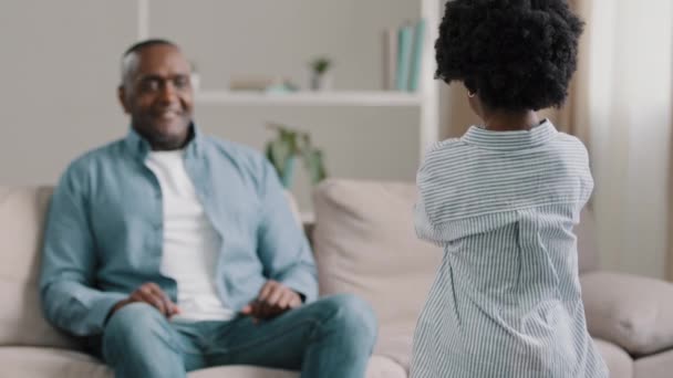 African american family mature dad sitting at home on couch surprised accepting birthday greetings little loving daughter gives handmade card for fathers day child comes to happy man hugs tightly — Video