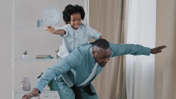 Mature african american grandpa plays with little granddaughter joyful kid pretends to be flying on plane sitting on grandfathers back funny male spread arms showing wings happy family enjoy playtime — Stockvideo