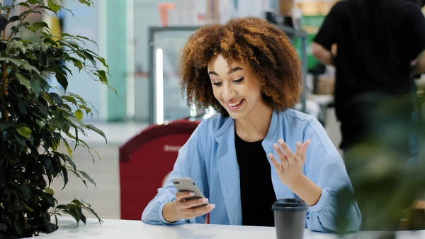 Jeune belle fille adolescente millénariste afro-américaine avec les cheveux bouclés se trouve dans le café en regardant le téléphone mobile se réjouit gagne obtient la victoire bon résultat gagner dans le succès du jeu en ligne chance triomphe — Photo