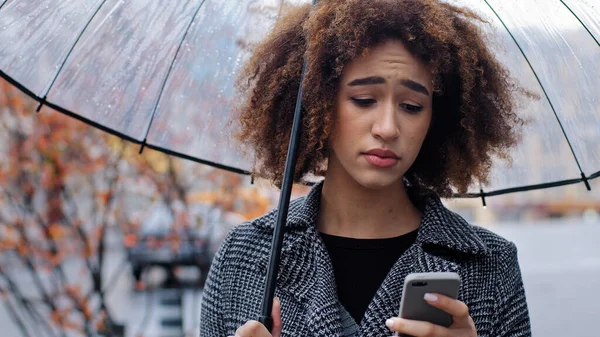 Афроамериканка кудрявая девушка с прозрачным зонтиком стоит на осенней улице под дождем смотрит на мобильный телефон расстроен грусть беспокоясь о низких батареях проблемы с интернетом плохие новости отвергаются — стоковое фото