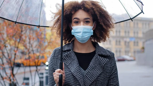 Красивая молодая афроамериканка с вьющимися волосами с прозрачным зонтиком стоит в городе на улице осенью холодный дождь носит медицинскую защитную маску от пандемии коронавируса — стоковое фото
