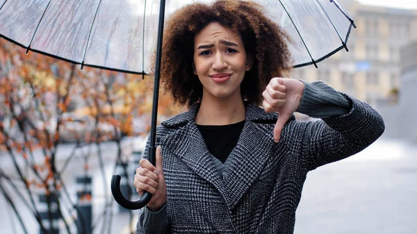 African american sad girl woman with curly hair with transparent umbrella shows gesture of disapproval refusal disagreement thumb down negative reaction no answer standing in city in rain bad weather