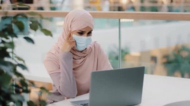 Pensive young muslim woman in medical mask in public place sitting typing on laptop arab girl in hijab writer journalist businesswoman student thinking deep in thought looking away found solution idea