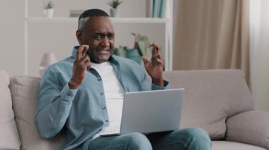 Mature african american man sitting in room fingers crossed hopefully looking at laptop screen asks good luck hopes to win male rejoices celebrating victory clapping hands makes gesture of approval