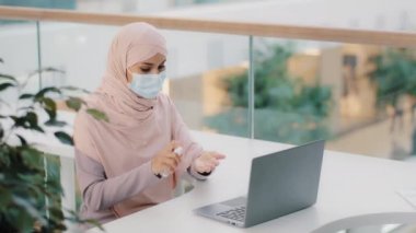 Koruyucu tıbbi maske takan genç Müslüman kadın antiseptik İslamcı kız yöneticiyi halka açık bir masada oturtup avuç içi bilgisayarlarını antibakteriyel ilaçla siliyor.