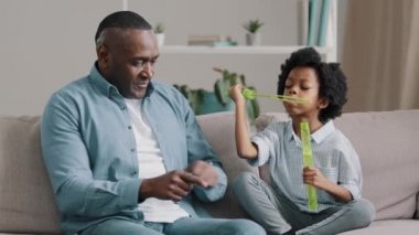 Adult african american grandfather with granddaughter sitting on sofa in room playing having fun together little cute girl blowing bubbles smiling mature man actively moves hands bursts soap balls