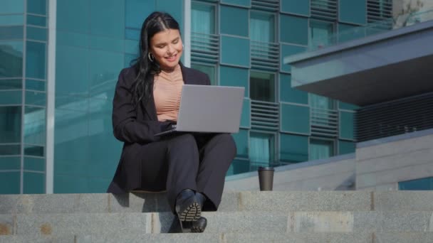 Young happy businesswoman spends leisure sitting outdoors on city building background joyful hispanic girl in business suit talking friendly by video call via webcam on laptop shows new place of work — 图库视频影像