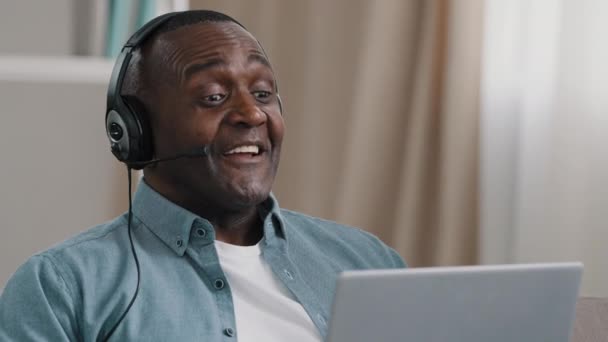 Close-up adult african american man in headphones communicating on webcam on laptop using video calling app hotline operator speaks into microphone uses headset male conducts webinar during quarantine — Wideo stockowe